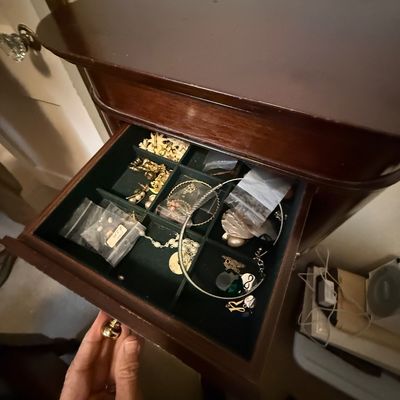 Open top drawer of a mahogany jewelry armoire, filled with various gold-tone costume jewelry, necklaces, and small accessories in...