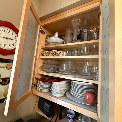Open light wood cabinet with etched glass doors filled with glassware, dishware, and decorative serving pieces.