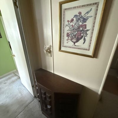 Framed floral print in a gold-toned frame hanging above a small dark wood corner cabinet with paneled doors.