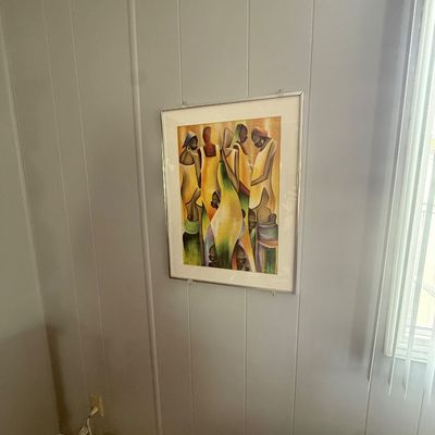 Framed African art print depicting stylized figures in yellow and earth-toned garments, mounted under glass in a metal frame.