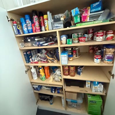 Tall wooden pantry cabinet filled with various household items, canned goods, boxed foods, cleaning supplies, and storage bags.