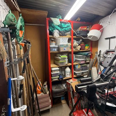 Workshop shed filled with long-handled yard tools, red shelving units with organized storage bins, and various power tools.