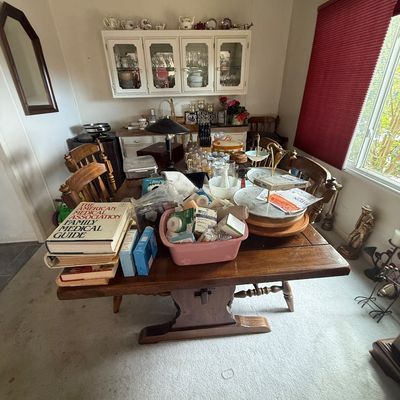 A wooden trestle dining room table loaded with various household items, including medical books, a pink tub filled with supplies,...