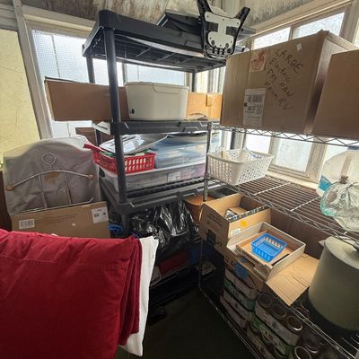 Assorted household goods, storage plastic shelving, metal wire racks, and cardboard boxes stacked in a porch area.