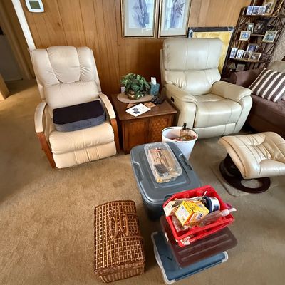 Pair of cream leather armchairs accompanied by a wooden end table, matching ottoman, and various storage bins in a wood-paneled room.