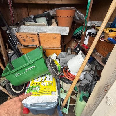 A storage shed filled with various lawn and garden tools, including a Scotts AccuGreen walk-behind fertilizer spreader, bags of NuLife...