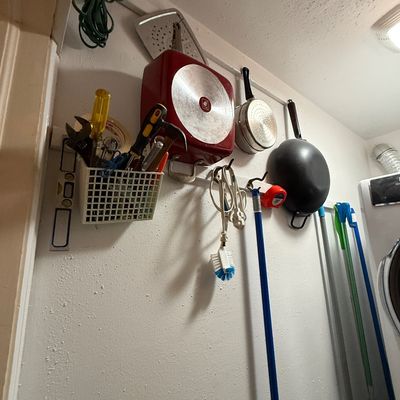 Assorted household tools, cookware, and cleaning supplies mounted on wall hooks and a utility rack, including a red square skillet, a...