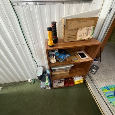 Small wood-grain laminate bookcase holding flashlights, a Pampered Chef cardboard box, small ceramic knickknacks, and various household...