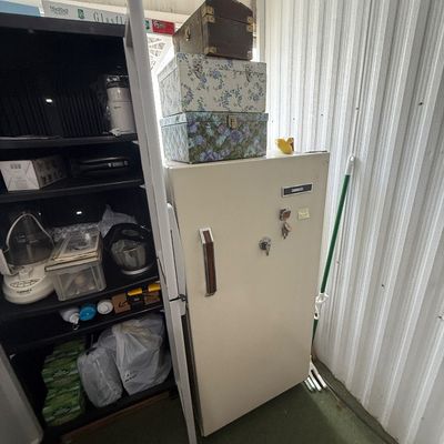 Vintage white metal safe standing next to a black shelving unit filled with small kitchen appliances and household items, topped with...
