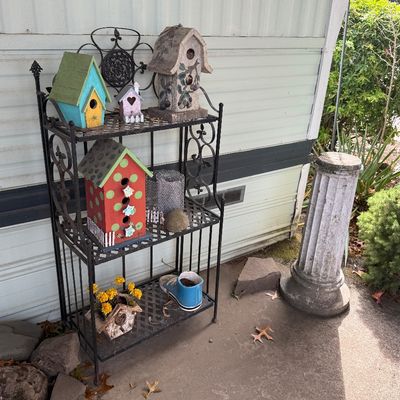 Black wrought iron three-tier baker's rack holding various colorful decorative birdhouses and garden accents, displayed alongside a...