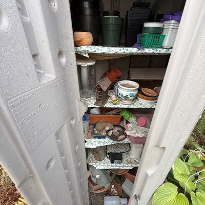 Plastic outdoor storage cabinet filled with an assortment of gardening supplies, plastic and ceramic plant pots, and lawn decor.
