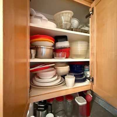 Open kitchen cabinet filled with assorted plastic food containers, mixing bowls, small appliances, and dishware.