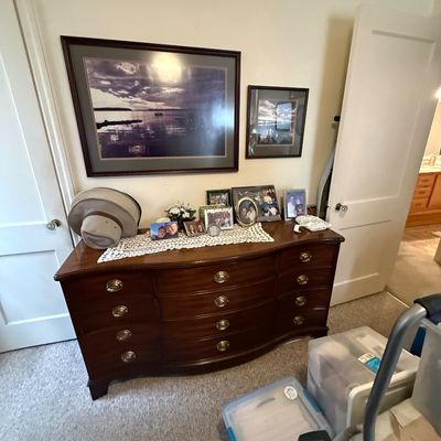 Traditional mahogany serpentine-front dresser with brass bail handles, displayed with framed artwork, family photos, and a sun hat.