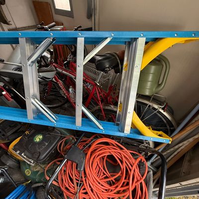 Blue fiberglass extension ladder stored in a shed alongside bicycles, lawncare equipment, and an orange extension cord.