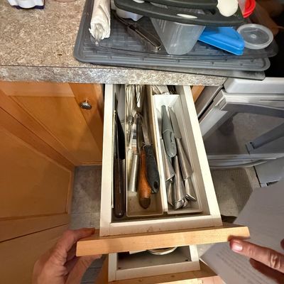 Open kitchen drawer filled with an assortment of utensils, including knives, a whisk, and flatware, organized in plastic tray inserts.