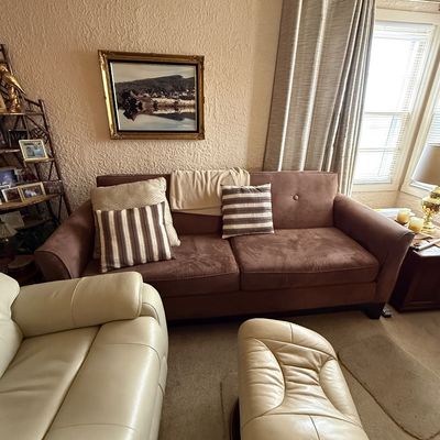 Brown microfiber three-seater sofa styled with throw pillows, alongside an off-white leather recliner armchair and matching swivel ottoman.