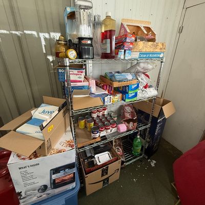 Four-tier wire shelving unit filled with household goods, canned food, small appliances, and office supplies alongside boxed items.
