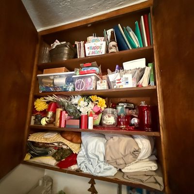 Open wooden wall cabinet filled with various household items, including books, craft supplies, artificial flowers, glass jars, and folded...