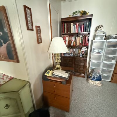 Home office setup featuring a wooden three-drawer filing cabinet topped with a brass lamp, a tall dark wood bookcase filled with books,...