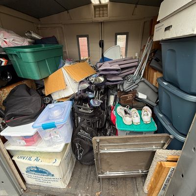 An outdoor storage shed filled with various household items, plastic storage totes, golf clubs, shoes, and folding chairs.