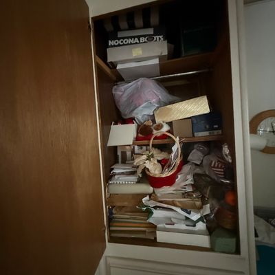 A built-in closet cabinet packed with various household goods, stationery, decorative items, and shoe boxes including a Nocona Boots box.