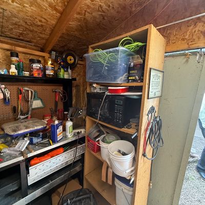 Workshop storage area featuring a tall wooden shelving unit packed with tools, extension cords, buckets, and a Black & Decker microwave.