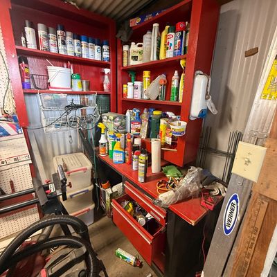 Workshop storage setup featuring red wooden shelving units filled with various household chemicals, paints, and lawn care supplies...