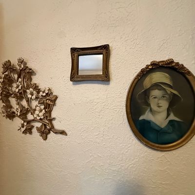 Collection of vintage wall decor featuring an oval framed child portrait, a small ornate gilt-framed mirror, and a floral branch wall...