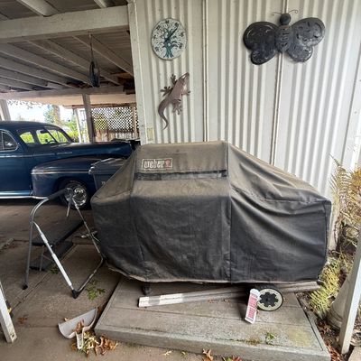 Covered Weber barbecue grill sitting on a wooden platform in a carport alongside wall decor and a folding step ladder.