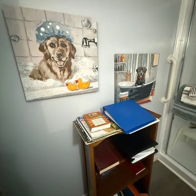 Pair of whimsical canvas wall art pieces featuring dogs taking baths, hanging above a small wooden bookcase stacked with books and binders.