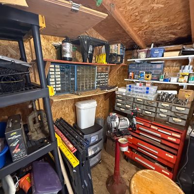 Workshop shed filled with tools and hardware, featuring a red bench grinder on a pedestal, rolling metal tool chests, plastic storage...