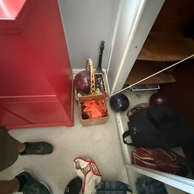 Collection of miscellaneous household items on carpeted floor, including bowling balls, carrying bags, a small cardboard box, and a red...