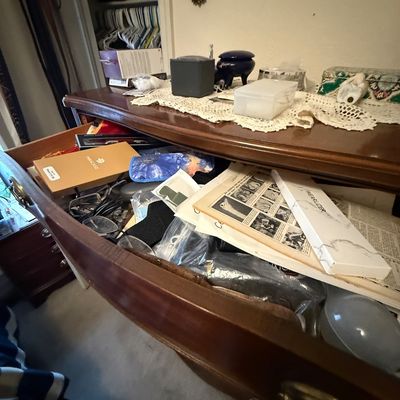 Open top drawer of a traditional dark wood dresser filled with miscellaneous items including boxed watch bands, eyeglasses, and printed...