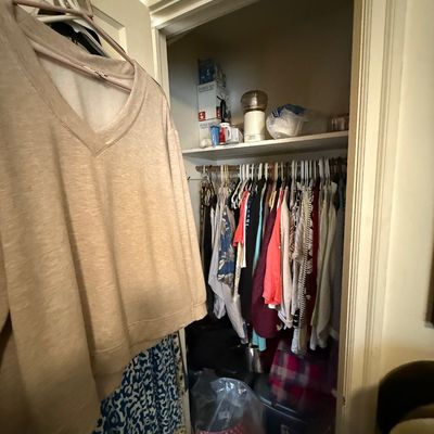 A closet filled with women's clothing on hangers, small appliances on the top shelf, and storage bins below, with a beige sweater...