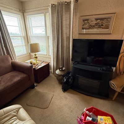 Living room setup featuring a flat-screen TV on an electric fireplace media stand, a brown plush armchair, a wood end table with a brass...