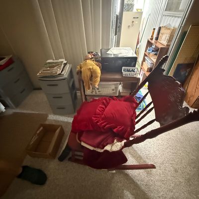 Vintage wooden rocking chair with carved crest rail holding red throw pillows, surrounded by household storage drawers, a doll, and a...