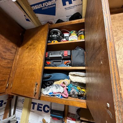 Open wooden wall cabinet filled with various workshop tools, hardware, power tools, paint cans, and cleaning supplies.