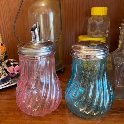 Sale Photo Thumbnail #91: Pair of vintage swirl glass sugar dispensers in pink and blue with metal lids, displayed alongside other glassware.