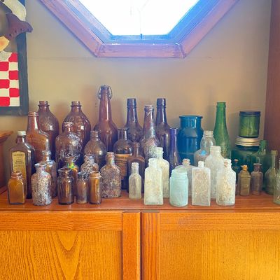 Sale Photo Thumbnail #4: Collection of antique glass bottles and apothecary jars in various shapes, sizes, and colors displayed on a wooden cabinet top.