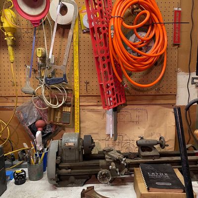 Sale Photo Thumbnail #106: Vintage Craftsman metal lathe set on a workbench, surrounded by assorted workshop tools, an orange air hose hanging on a pegboard, and a wall-mounted wooden barometer.