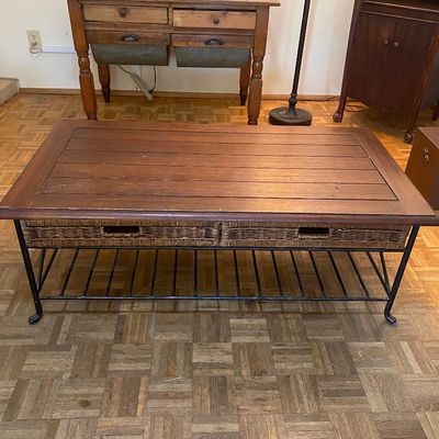 Sale Photo Thumbnail #29: Rustic wood and metal coffee table featuring a planked tabletop, woven wicker drawer fronts, and a lower slatted metal shelf. Behind it stands a vintage wooden baker's table with flour bins and a dark wood cabinet.