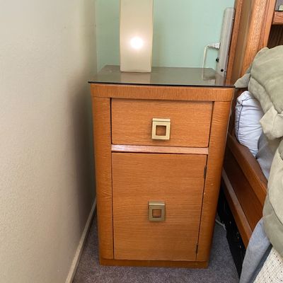 Sale Photo Thumbnail #83: Mid-century oak nightstand featuring square brass hardware, a top drawer, and a lower cabinet door, topped with a small rectangular table lamp.