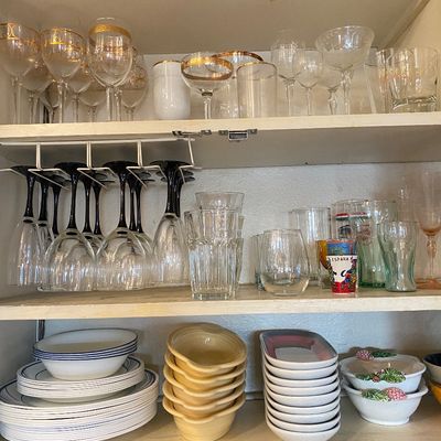 Sale Photo Thumbnail #48: Cabinet shelves organized with various glassware including gold-rimmed goblets, black-stemmed wine glasses, and etched crystal, above stacks of ceramic dishes and bowls.