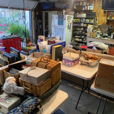 Sale Photo Thumbnail #111: A garage packed with estate sale items arranged on folding tables, including woven picnic baskets, plush toys, framed artwork, and household goods.
