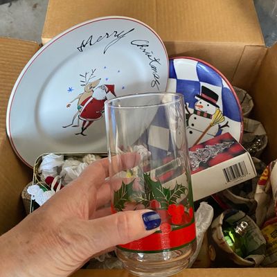 Sale Photo Thumbnail #121: Cardboard box packed with holiday tableware and decor, featuring a holly-patterned drinking glass, Santa and snowman decorative plates, and various boxed ornaments.