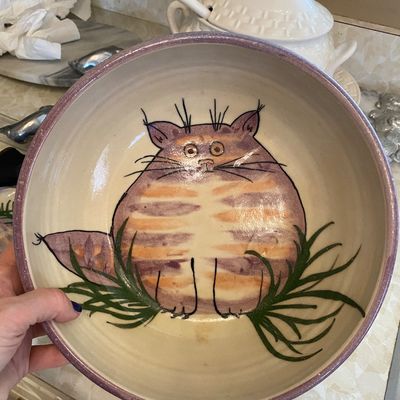 Sale Photo Thumbnail #74: Handcrafted studio pottery bowl featuring a whimsical, hand-painted striped cat motif framed by green foliage with a purple-rimmed edge.