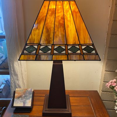 Sale Photo Thumbnail #35: Mission-style stained glass table lamp with a dark metal tapered base and amber and green slag glass shade, illuminated on a wooden side table.