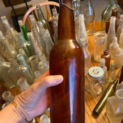 Sale Photo Thumbnail #53: Vintage amber glass lightning stopper bottle held by a hand, part of a large collection of vintage glass bottles and jars displayed on a wooden table.
