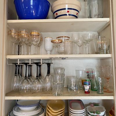 Sale Photo Thumbnail #11: Assorted vintage kitchenware and glassware arranged inside a white painted cupboard. Includes gold-rimmed wine glasses, black-stemmed goblets, clear drinking glasses, stoneware bowls, and stacked dinnerware.
