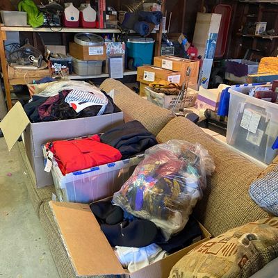 Sale Photo Thumbnail #112: A garage sale setup featuring a brown textured sofa loaded with boxes and bags of clothing, plastic storage bins, and various household goods.